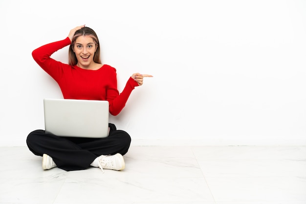 Jovem mulher caucasiana com um laptop sentada no chão surpresa e apontando o dedo para o lado