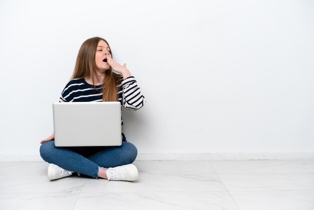 Jovem mulher caucasiana com um laptop sentado no chão isolado no fundo branco, bocejando e cobrindo a boca aberta com a mão