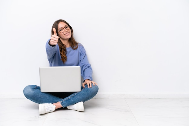 Jovem mulher caucasiana com um laptop sentado no chão isolado no fundo branco com polegares para cima porque algo bom aconteceu