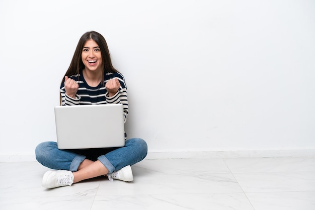 Jovem mulher caucasiana com um laptop sentado no chão isolado no fundo branco comemorando uma vitória na posição de vencedor