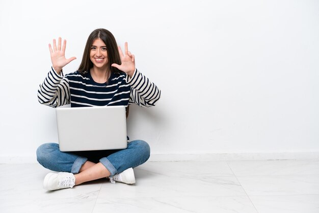 Jovem mulher caucasiana com um laptop sentado no chão isolado no fundo branco contando oito com os dedos