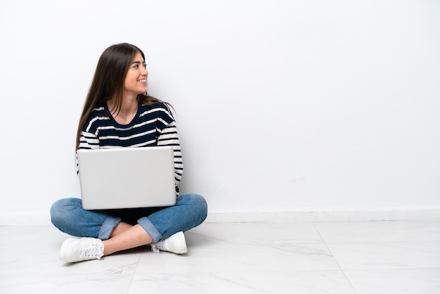 Jovem mulher caucasiana com um laptop sentado no chão isolado no fundo branco em posição lateral