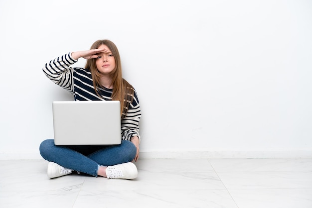 Jovem mulher caucasiana com um laptop sentado no chão isolado no fundo branco, olhando para longe com a mão para procurar algo