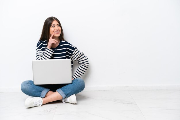 Jovem mulher caucasiana com um laptop sentado no chão isolado no fundo branco pensando em uma ideia enquanto olha para cima