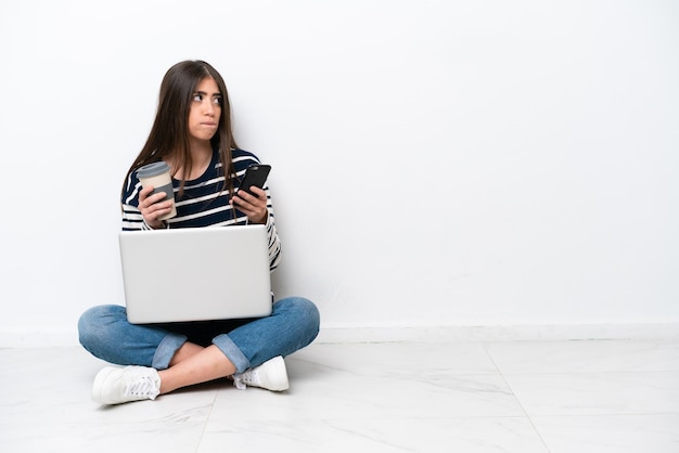 Jovem mulher caucasiana com um laptop sentado no chão isolado no fundo branco segurando café para levar e um celular enquanto pensa em algo