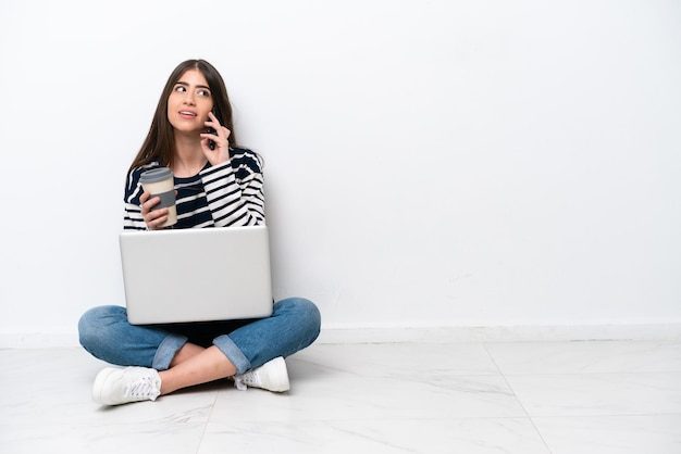 Jovem mulher caucasiana com um laptop sentado no chão isolado no fundo branco segurando café para levar e um celular