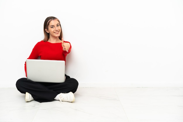 Jovem mulher caucasiana com um laptop sentado no chão, mostrando e levantando um dedo