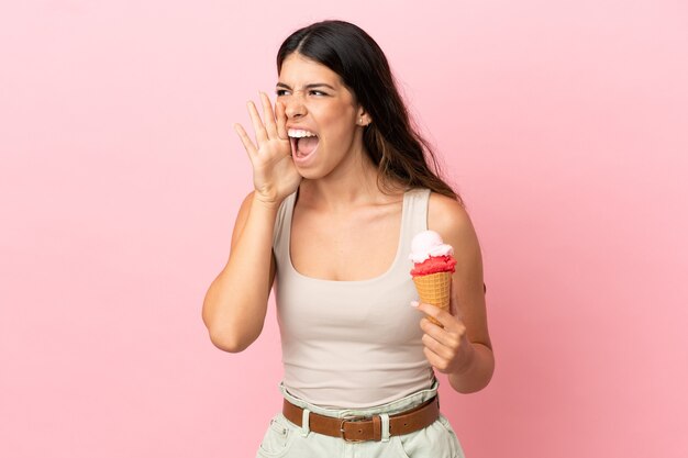 Jovem mulher caucasiana com um sorvete de corneta isolado em um fundo rosa gritando com a boca bem aberta para o lado