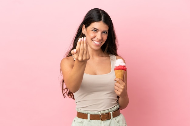 Jovem mulher caucasiana com um sorvete de corneta isolado no fundo rosa, convidando para vir com a mão. Feliz que você veio