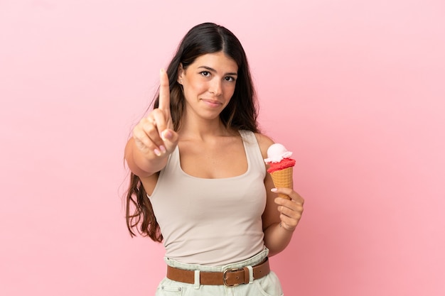 Jovem mulher caucasiana com um sorvete de corneta isolado no fundo rosa, mostrando e levantando um dedo