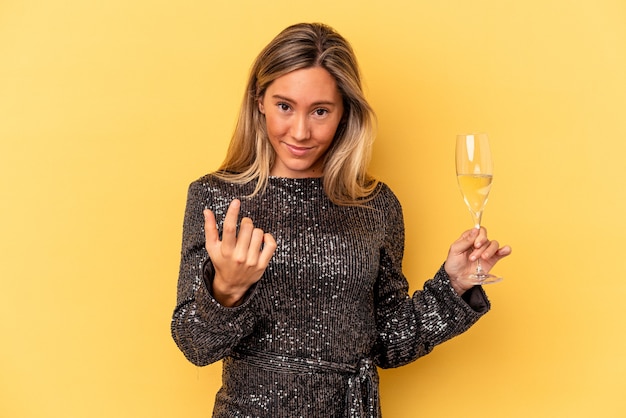 Jovem mulher caucasiana, comemorando o ano novo segurando a taça de cava isolada no fundo amarelo, apontando com o dedo para você como se estivesse convidando a se aproximar.