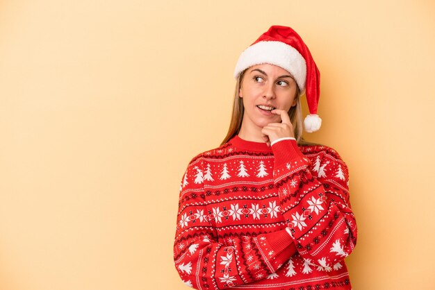 Jovem mulher caucasiana, comemorando o Natal, isolado em um fundo amarelo relaxado pensando em algo olhando para um espaço de cópia.