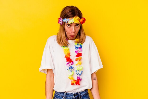 Jovem mulher caucasiana, comemorando uma festa havaiana isolada em amarelo encolhe os ombros e abre os olhos confusos.