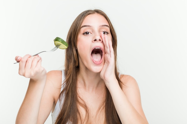 Jovem mulher caucasiana comendo pepino gritando animado para a frente.