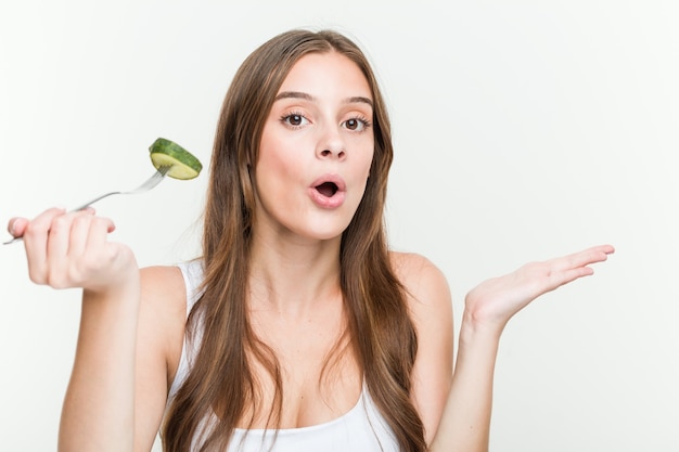 Jovem mulher caucasiana comendo pepino impressionada, segurando o espaço da cópia na palma da mão.