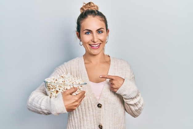 Jovem mulher caucasiana comendo pipoca sorrindo feliz apontando com a mão e o dedo