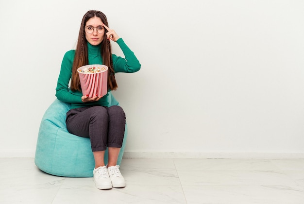 Jovem mulher caucasiana comendo pipocas em um sopro isolado no fundo branco, apontando o templo com o dedo, pensando, focado em uma tarefa.