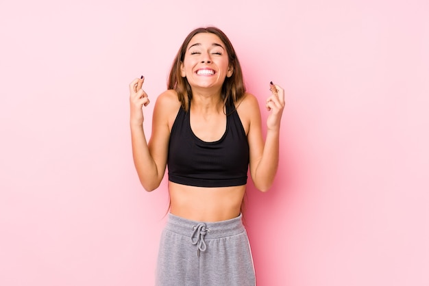 Jovem mulher caucasiana de fitness posando em uma parede rosa cruzando os dedos para ter sorte
