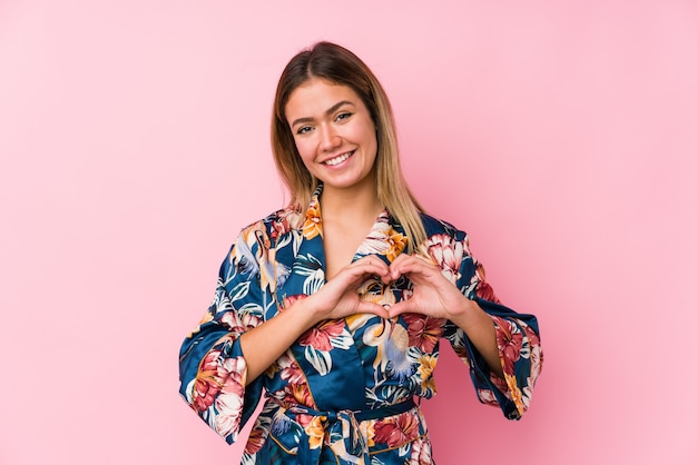 Jovem mulher caucasiana de pijama, sorrindo e mostrando uma forma de coração com as mãos.