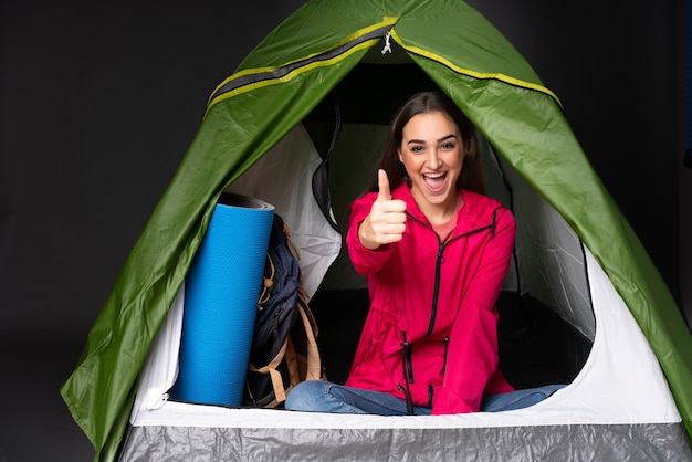 Jovem mulher caucasiana dentro de uma barraca de acampamento verde com o polegar para cima porque algo bom aconteceu