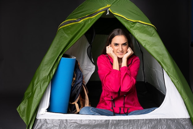 Foto jovem mulher caucasiana dentro de uma barraca verde de acampamento frustrada e cobrindo as orelhas