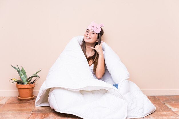 Jovem mulher caucasiana, descansando com uma colcha, mostrando um gesto de chamada de celular com os dedos.