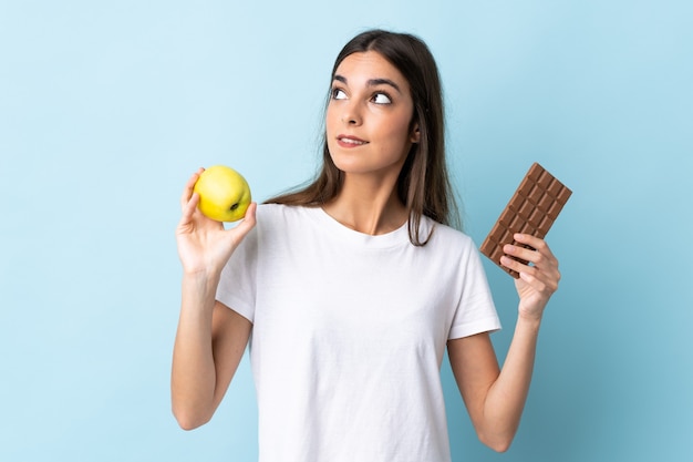 Jovem mulher caucasiana em azul tendo dúvidas enquanto toma um comprimido de chocolate com uma das mãos