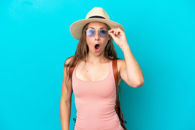 Jovem mulher caucasiana em férias de verão isolada em um fundo azul com óculos e surpresa