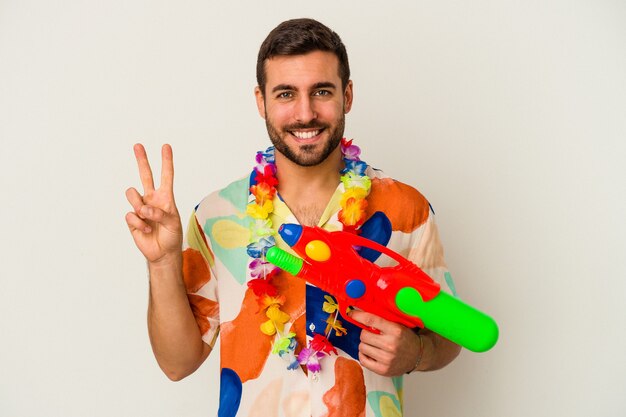 Jovem mulher caucasiana em uma festa havaiana segurando uma pistola de água isolada na parede branca, mostrando o número dois com os dedos.