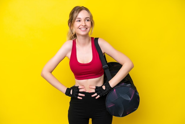 Jovem mulher caucasiana esportiva com fundo isolado de bolsa esportiva posando com os braços no quadril e sorrindo