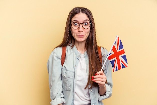 Jovem mulher caucasiana estudando inglês isolada em fundo amarelo encolhe os ombros e abre os olhos confusos