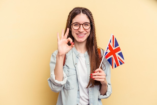 Jovem mulher caucasiana estudando inglês isolado em fundo amarelo alegre e confiante, mostrando o gesto okey