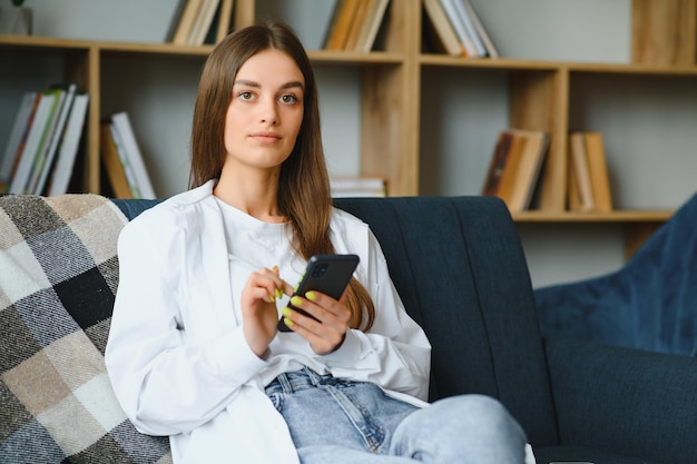 Jovem mulher caucasiana feliz, ela tem 20 anos em roupas casuais, segura na mão, usa celular, sente-se no sofá azul, descanso interno em casa, no próprio apartamento, conceito de estilo de vida de pessoas