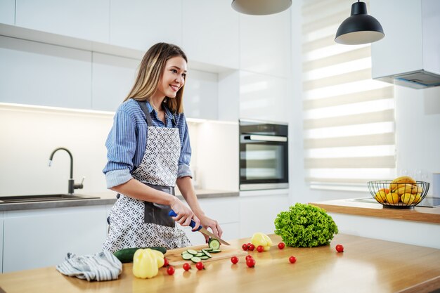 Jovem mulher caucasiana feliz no avental em pé na cozinha e cortar o pepino. No balcão da cozinha existem todos os tipos de vegetais. Conceito de alimentação saudável.