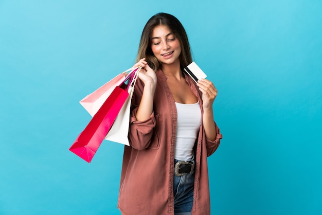 Jovem mulher caucasiana isolada em azul segurando sacolas de compras e um cartão de crédito