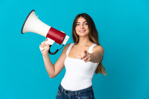 Jovem mulher caucasiana isolada em azul segurando um megafone e sorrindo enquanto aponta para a frente