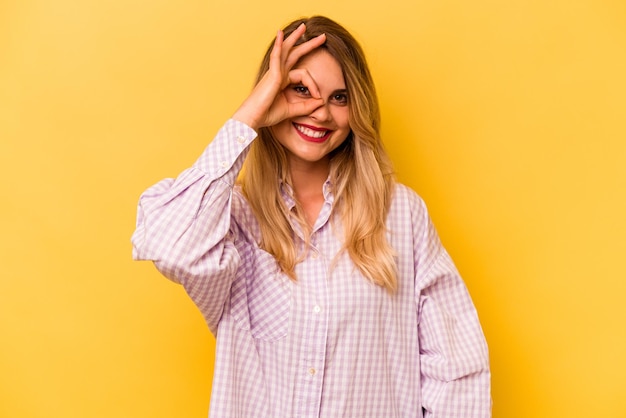 Jovem mulher caucasiana isolada em fundo amarelo animada mantendo o gesto ok no olho