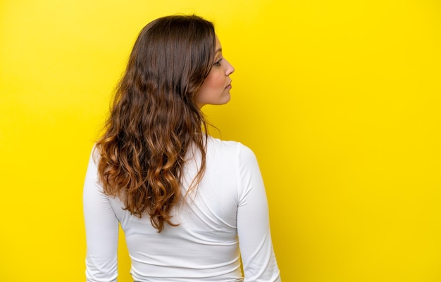 Jovem mulher caucasiana isolada em fundo amarelo em posição de costas e olhando para o lado