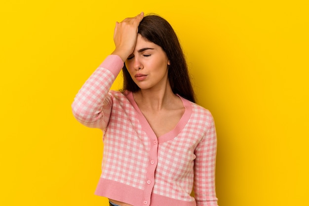 Jovem mulher caucasiana isolada em fundo amarelo, esquecendo de algo, batendo na testa com a palma da mão e fechando os olhos.