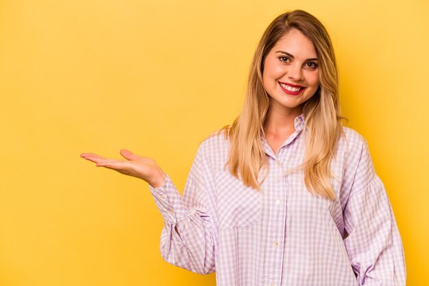 Jovem mulher caucasiana isolada em fundo amarelo, mostrando um espaço de cópia na palma da mão e segurando a outra mão na cintura
