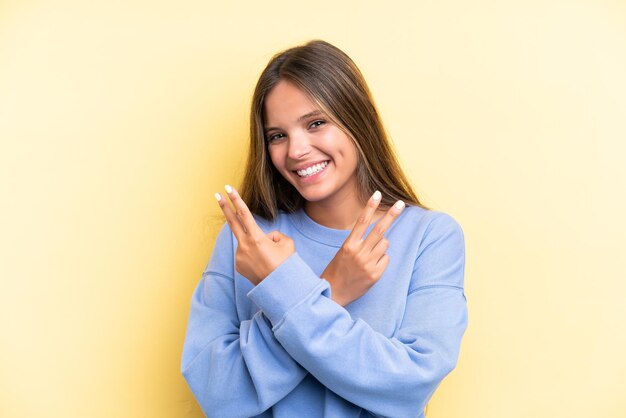 Jovem mulher caucasiana isolada em fundo amarelo sorrindo e mostrando sinal de vitória