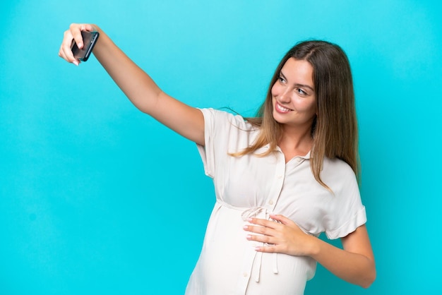 Jovem mulher caucasiana isolada em fundo azul grávida e fazendo uma selfie
