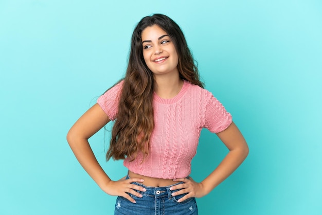 Jovem mulher caucasiana isolada em fundo azul posando com braços no quadril e sorrindo