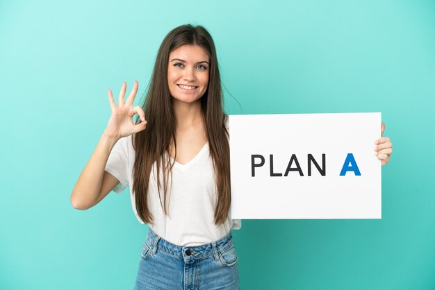 Jovem mulher caucasiana isolada em fundo azul segurando um cartaz com a mensagem PLANO A com sinal de ok