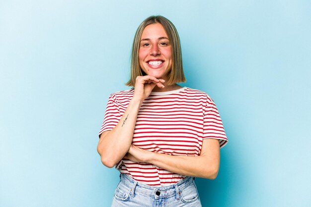 Jovem mulher caucasiana isolada em fundo azul sorrindo feliz e confiante tocando o queixo com a mão