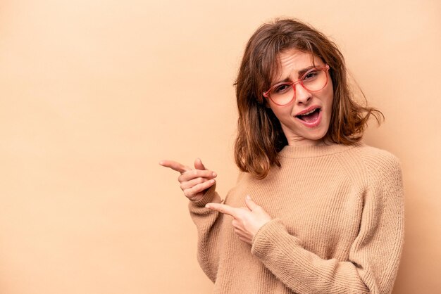 Jovem mulher caucasiana isolada em fundo bege apontando com os dedos indicadores para um espaço de cópia expressando emoção e desejo