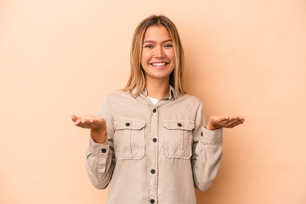 Jovem mulher caucasiana isolada em fundo bege faz escala com os braços, sente-se feliz e confiante.