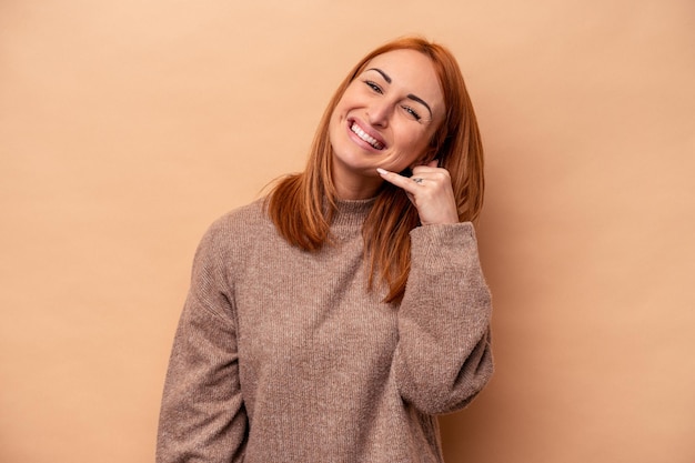 Jovem mulher caucasiana isolada em fundo bege, mostrando um gesto de chamada de celular com os dedos.