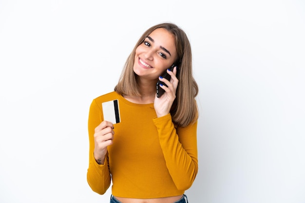 Jovem mulher caucasiana isolada em fundo branco, mantendo uma conversa com o celular e segurando um cartão de crédito