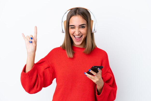 Jovem mulher caucasiana isolada em fundo branco ouvindo música com um celular fazendo gesto de rock